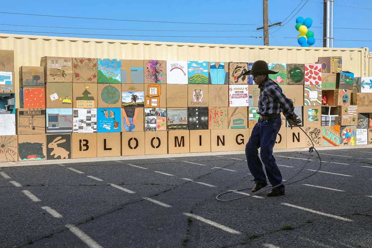 A boy swings a lasso in front of cardboard boxes decorated by community members as part of the “Bloomington Speaks” art installation.